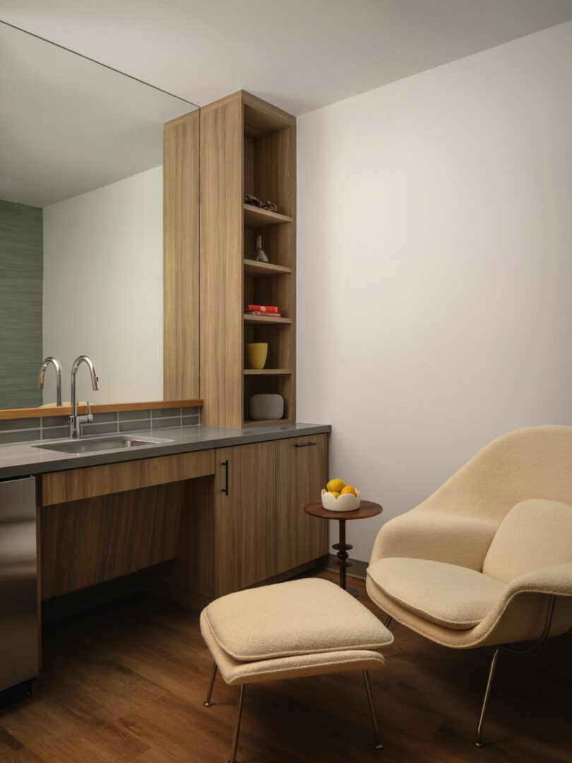 A minimalist kitchen area with a wooden cabinet, a sink, a shelf unit, and a beige armchair with an ottoman next to a small side table holding a bowl of lemons.