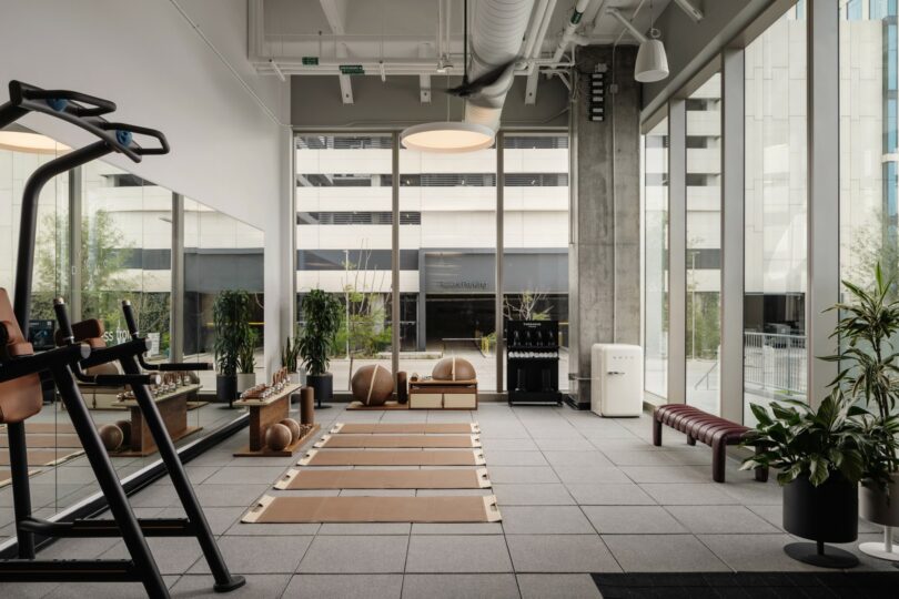 Modern gym with large windows, treadmills, exercise mats, exercise balls, and various equipment. Indoor plants and benches are placed around the space, providing a clean and minimalistic look.