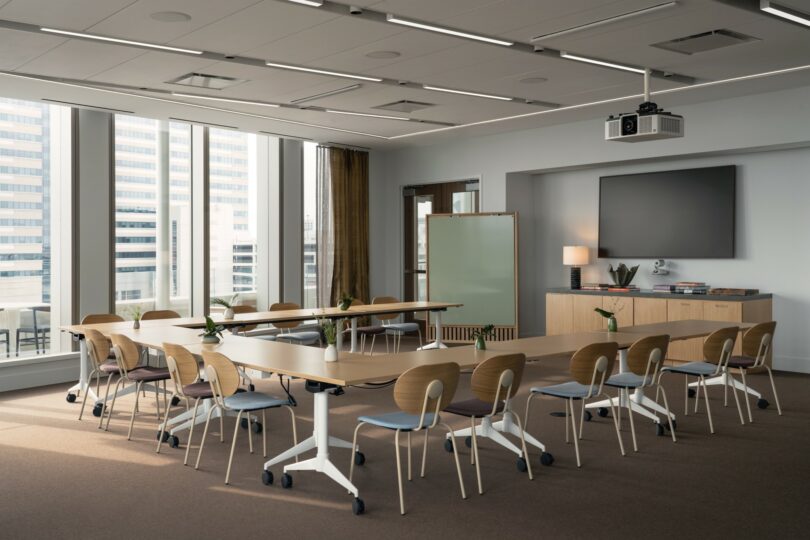 A modern conference room with U-shaped table setup, chairs, a large screen, a projector, and windows providing natural light.