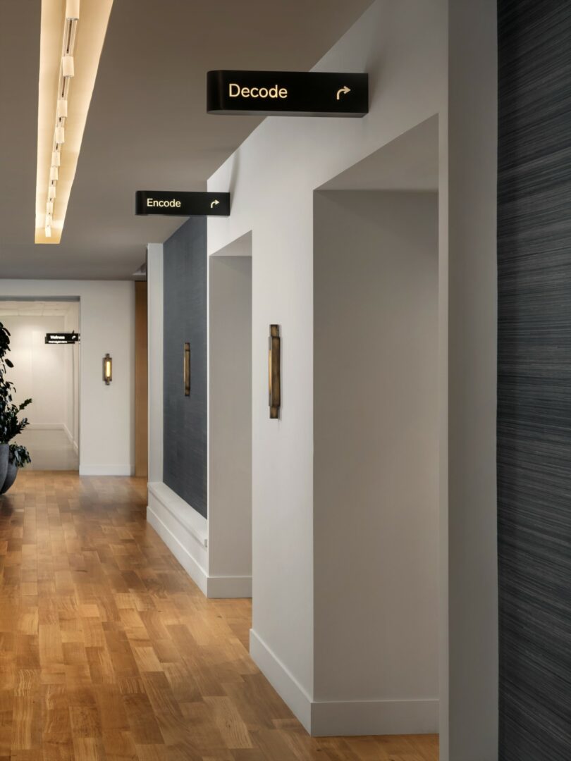 A hallway with wooden floors and white walls has signs indicating directions to rooms "Decode" and "Encode.