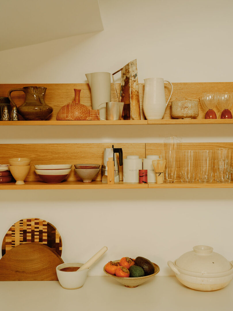 Kitchen shelves with assorted ceramics, glasses, and pitchers. Counter features wooden boards, a clay pot, a wooden spoon, and a bowl with tomatoes and avocados.