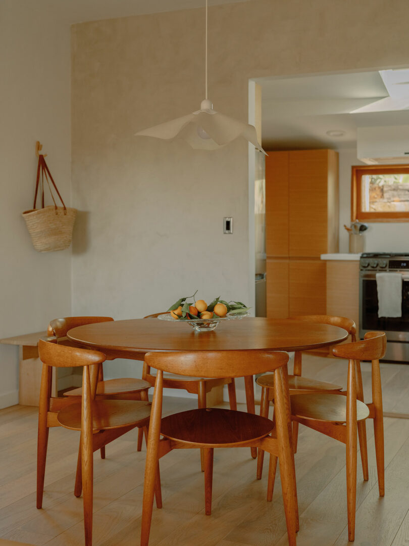 A wooden dining table with chairs, a bowl of fruit, and a hanging light fixture in a modern kitchen setting.