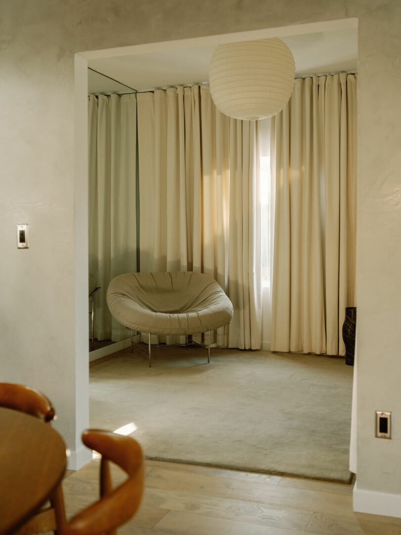 A cozy room with beige curtains, a soft round chair, a carpet, and a hanging paper lantern.