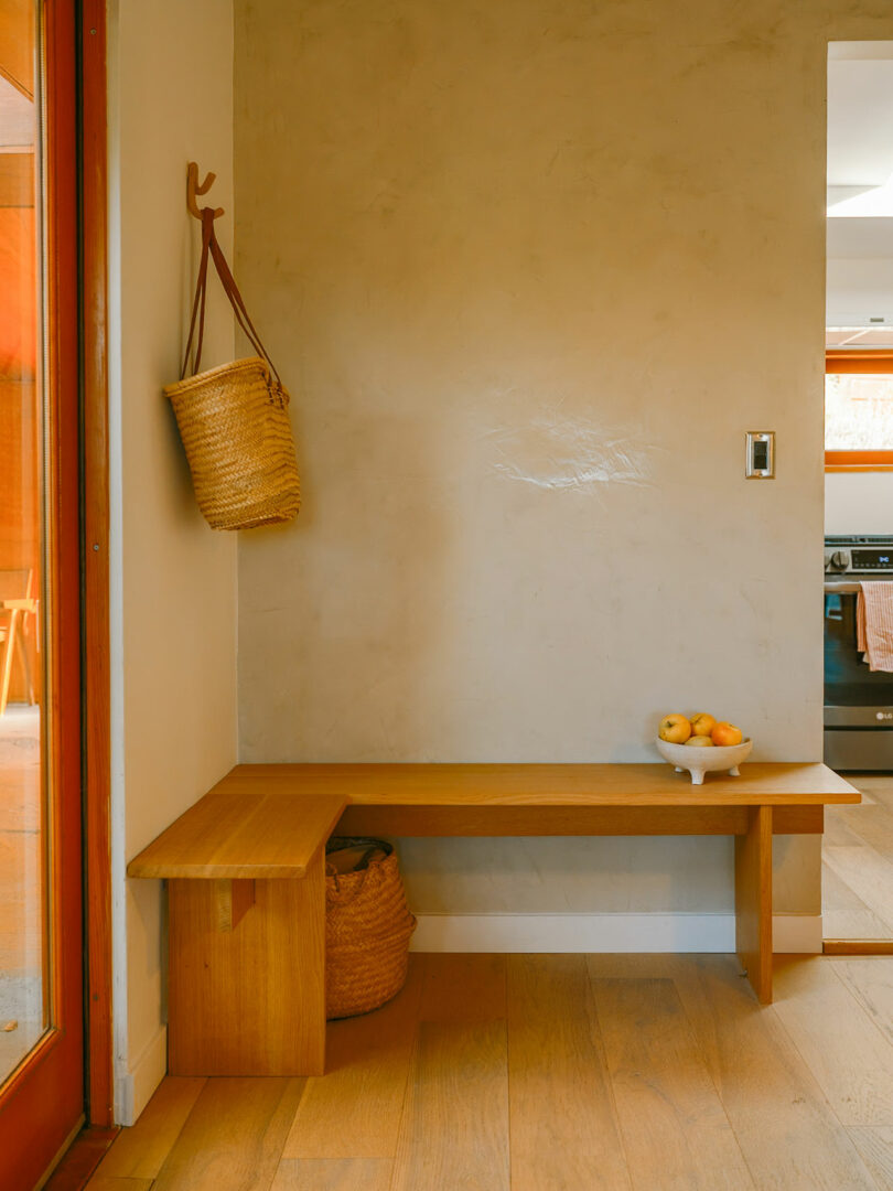 Minimalist entryway with a wooden bench, woven bag hanging on the wall, basket underneath, and a bowl of apples on the bench. A red door and a light switch are visible.