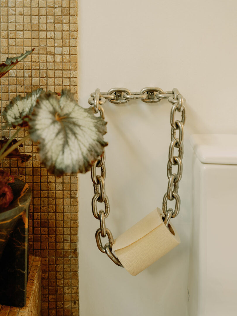 A roll of toilet paper is suspended on a metal chain attached to the wall next to a plant with large leaves. The background features a tiled wall.