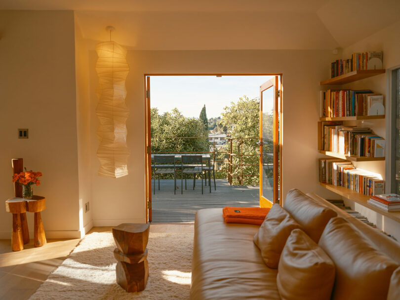 A cozy living room with a sofa, wooden decor, and bookshelves opens to a balcony with chairs, overlooking trees and distant structures. Warm lighting enhances the inviting atmosphere.