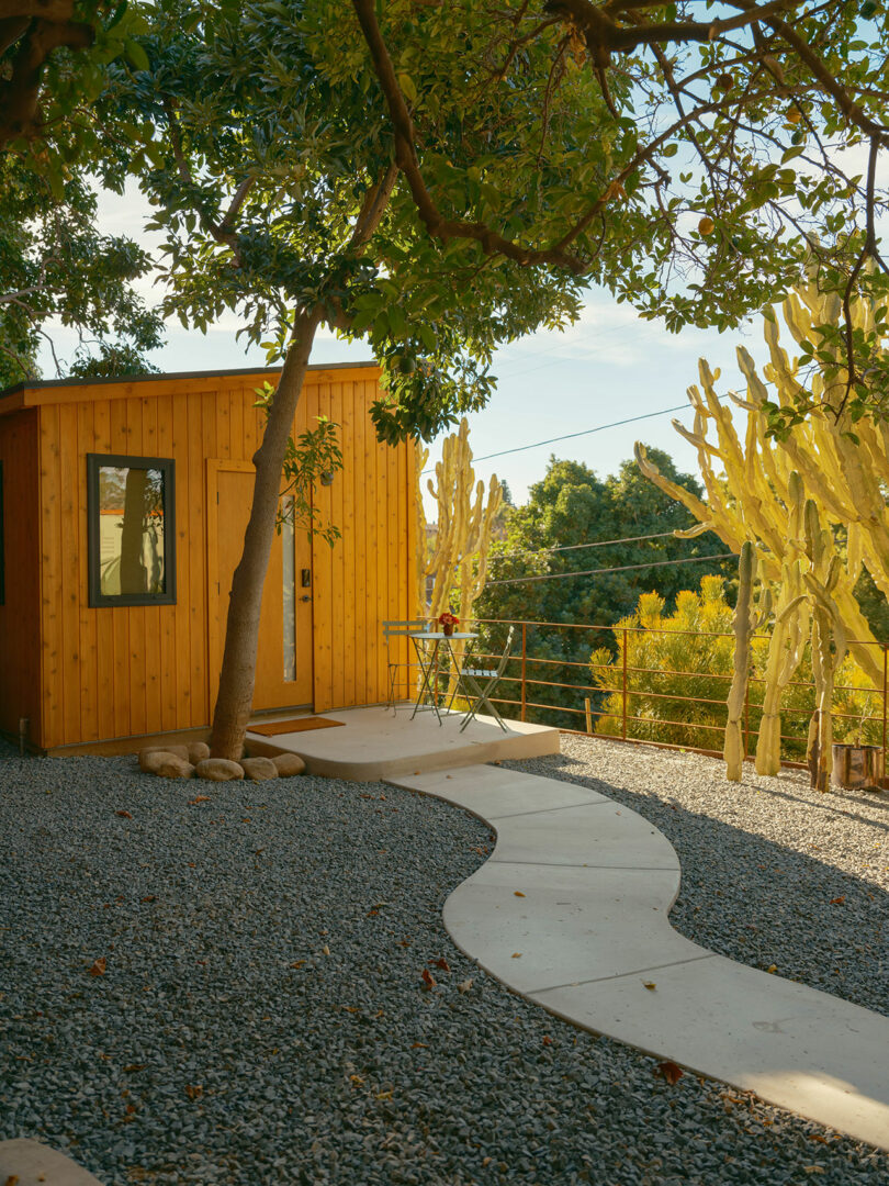 A small wooden cabin with a single window and door sits amidst desert plants, including tall cacti. A curved stone path leads to it, and a small table with chairs is nearby.