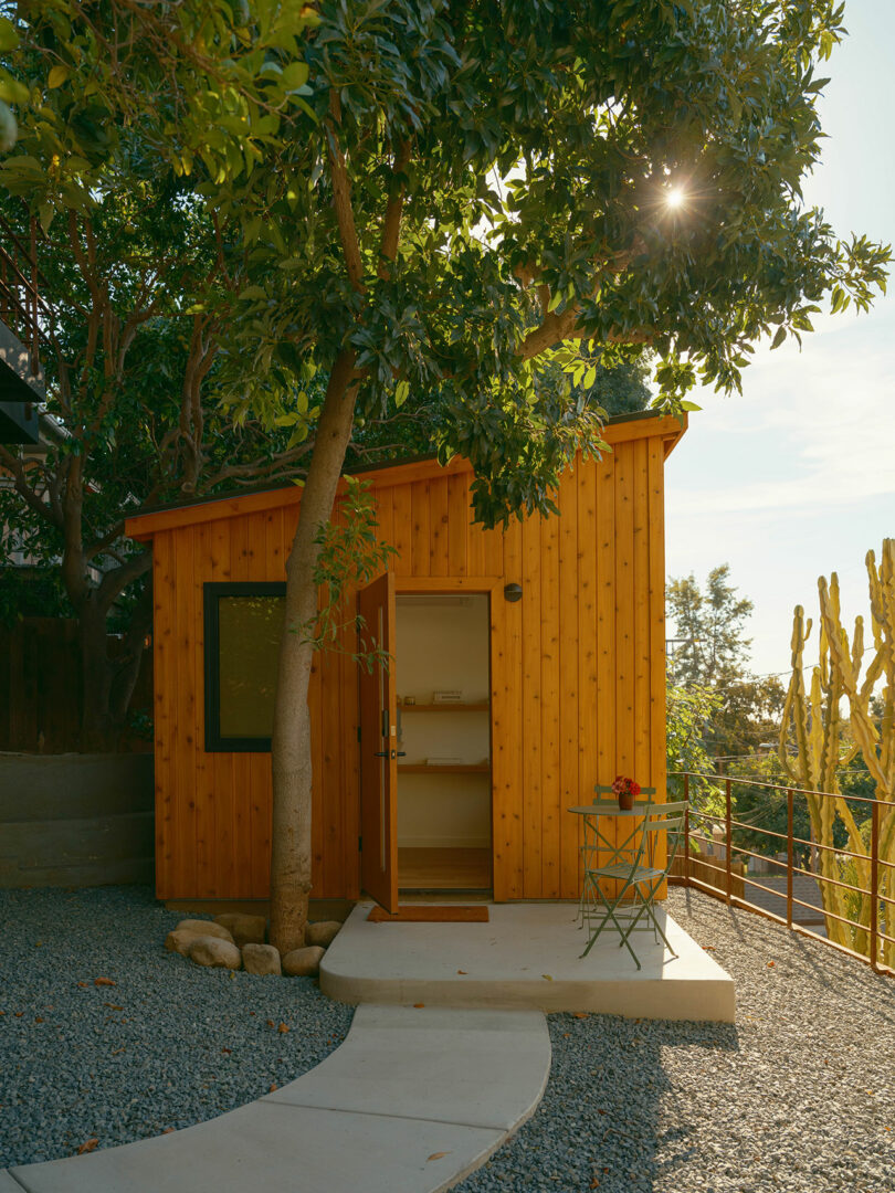 Small wooden cabin under a tree with a curved path. An open door reveals a glimpse inside. A round table with items sits outside. Sunlight filters through the branches.