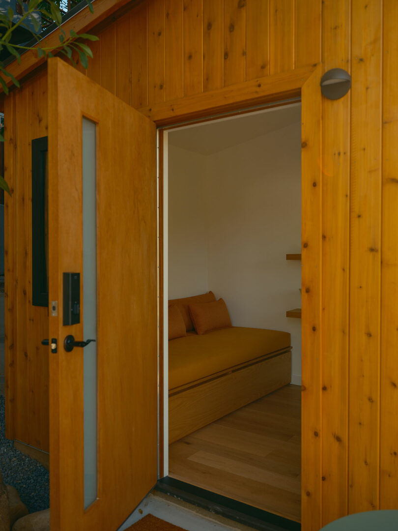 Open wooden door leading to a minimalist room with a bench and cushions inside a wooden building.