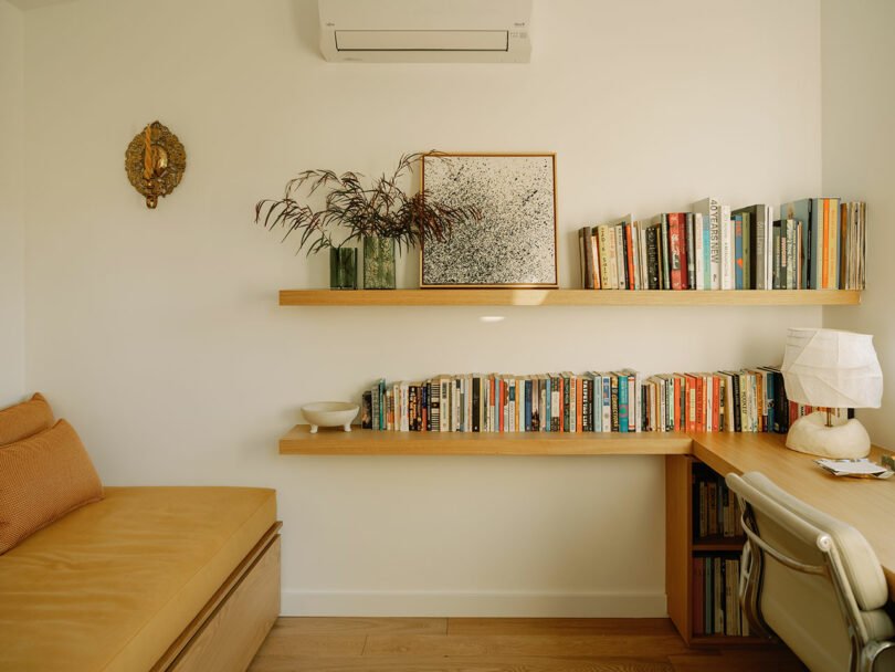 A cozy room with a beige sofa, bookshelves filled with books, a desk with a lamp, and a decorative wall hanging.