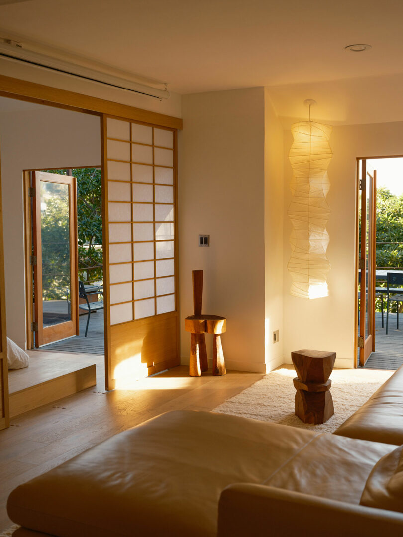 A sunlit room with wooden furniture, shoji screen doors, and textured paper lanterns, opening to a view of greenery outside.