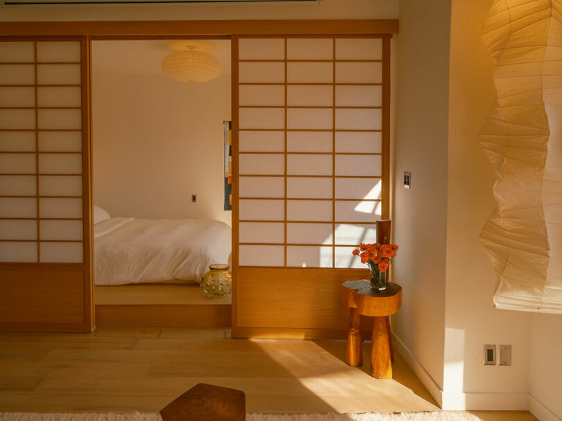 A serene bedroom with shoji sliding doors, a bed with white bedding, a small wooden side table holding orange flowers, and soft natural lighting.