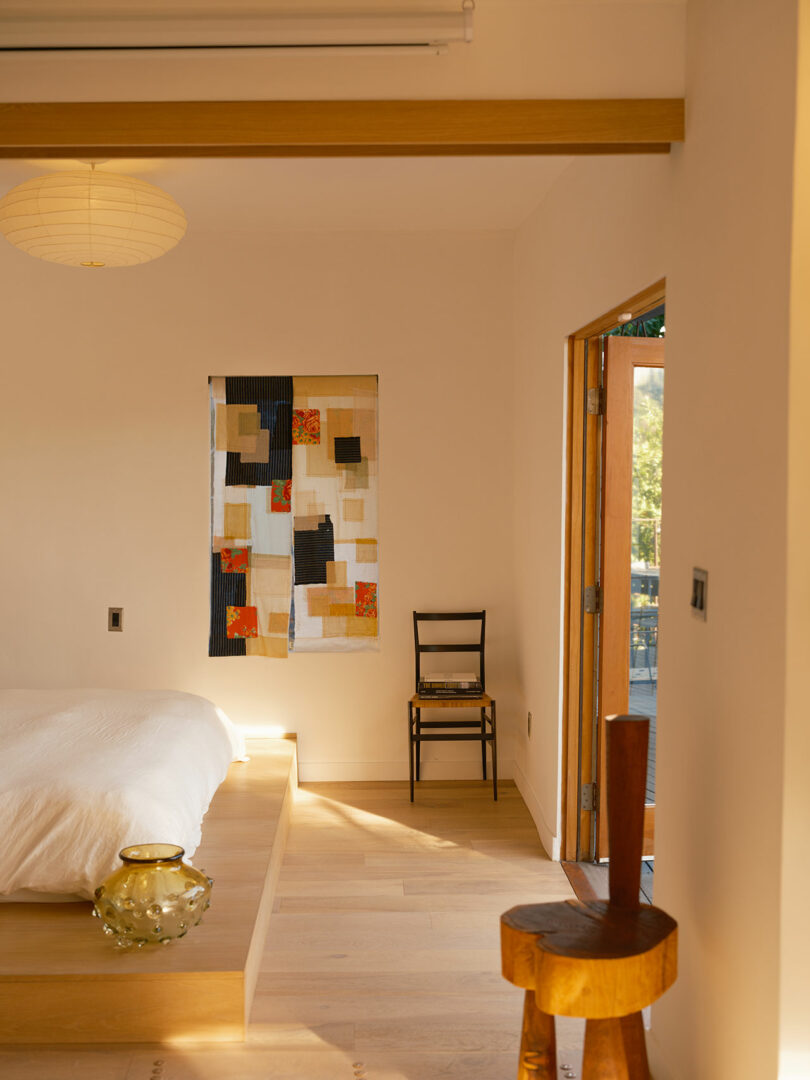 A minimalist bedroom with a platform bed, a patchwork wall hanging, wooden flooring, and natural light streaming through an open door. A wooden chair and a small round table are present.