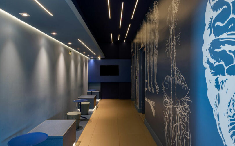 A dimly lit, modern hallway with blue walls, minimalist furniture from Estúdio Protobox, overhead lighting, and abstract white wall drawings. A flat-screen TV is mounted on the distant wall.