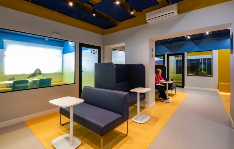 A modern office space features blue booths with white tables, glass-walled meeting rooms by Estúdio Protobox, and two people working at desks within the booths. Room numbers include T04 and T07.