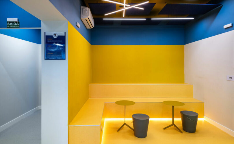 A modern, brightly lit waiting area designed by Estúdio Protobox features yellow tiered seating, two small round tables, and a blue and yellow color scheme. An emergency exit sign and an air conditioning unit are visible on the walls.