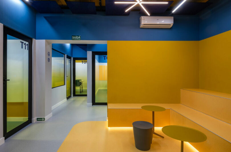 A modern, brightly lit room with blue and yellow walls, T11 and T05 marked on glass doors, tiered yellow seating, small round tables, and an air conditioning unit on the wall; this space is designed for both comfort and productivity.