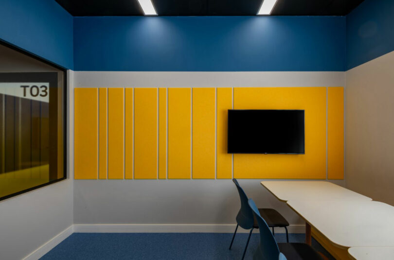 A small meeting room by Estúdio Protobox features blue walls, yellow acoustic panels, a wall-mounted TV, a white conference table, and blue chairs. A window labeled "T03" is on the left side.