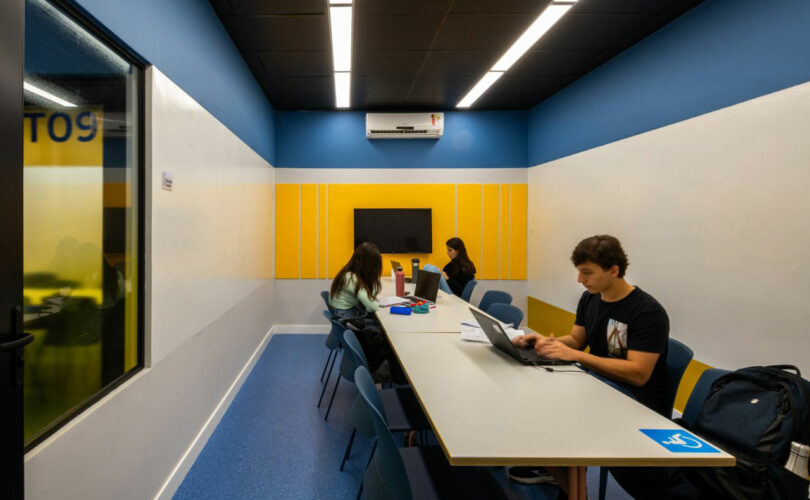 A group of people work on laptops at a long table in a brightly lit, modern study room with blue and yellow walls, reminiscent of the design aesthetics championed by Estúdio Protobox.