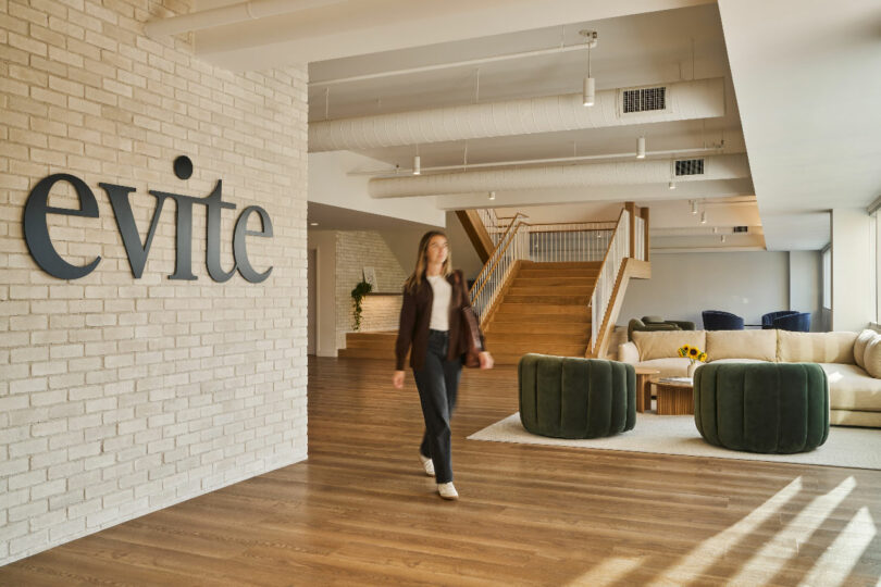 A woman walks through a modern office space designed by IA Interior Architects, with a sign reading "evite" on a white brick wall. A staircase, green cushioned chairs, and a beige couch are visible in the background.