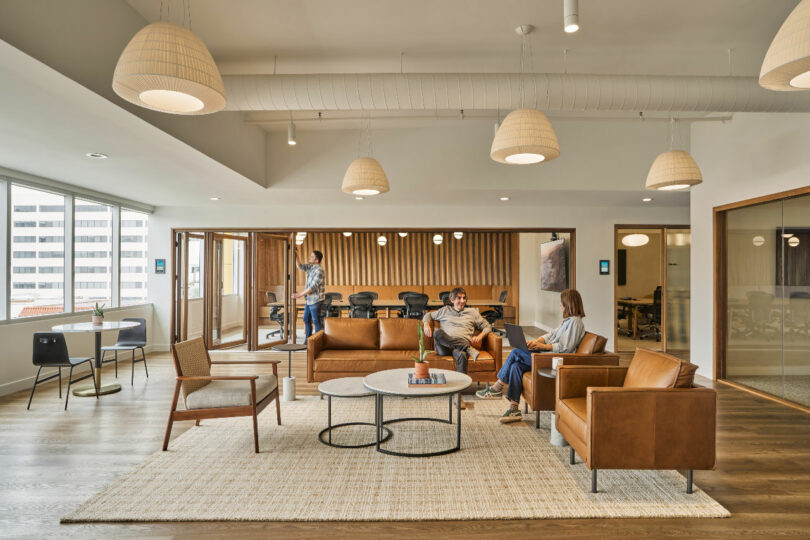 Modern office lounge area designed by IA Interior Architects, featuring leather sofas, chairs, coffee tables, and pendant lights. Three people are seated and conversing while another person moves in the background near a conference room.