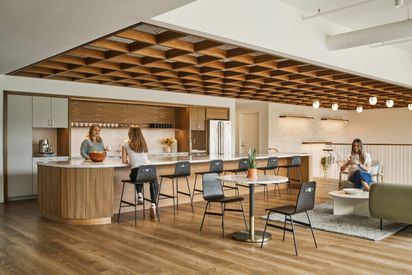 A modern open kitchen and lounge area designed by IA Interior Architects features two women talking at a counter while another woman sits on a couch, using a laptop. The space boasts wooden accents and contemporary furniture.