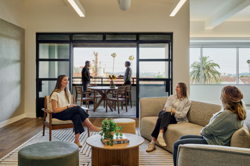 A group of people are sitting and talking in a modern office lounge with large windows, designed by IA Interior Architects. Two people are standing and conversing on a balcony outside. The space is decorated with plants and modern furniture.