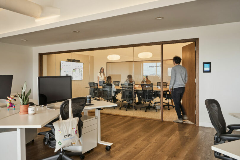 A modern office space designed by IA Interior Architects features several desks and computers in the foreground. Through a glass wall, a dynamic meeting room with people inside is visible, with one person entering the room.