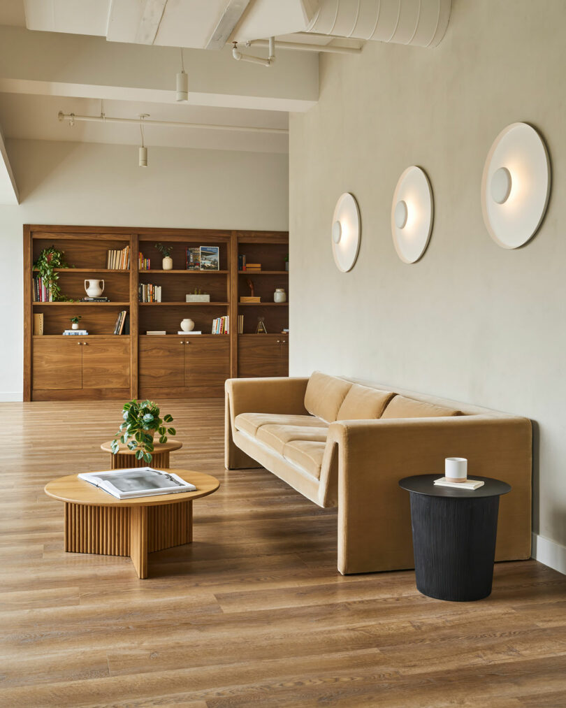 A modern living room designed by IA Interior Architects features a beige sofa, a wooden coffee table, a black side table, wall-mounted round lights, a plant, and a bookshelf with various items and books against a light-green wall.