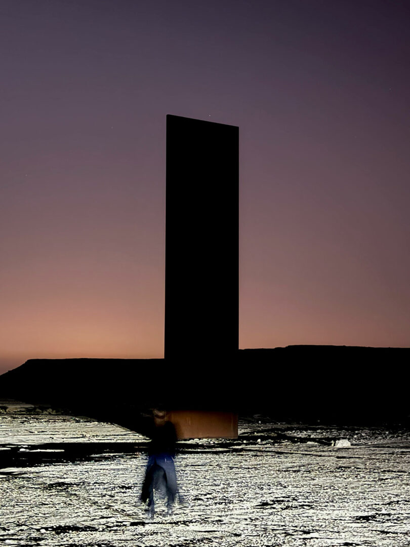 A person walks near a tall, dark monolith standing upright on a textured landscape at dusk or dawn.