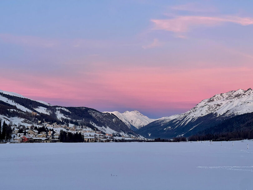 Snow-covered landscape with mountains in the background under a pink and blue sky at sunset. A small village is visible on the left side.