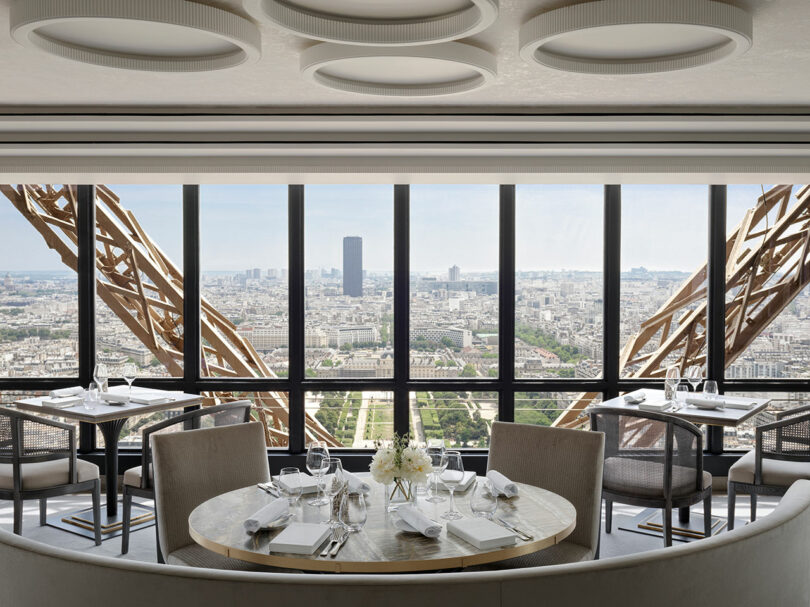 Restaurant interior with tables set for dining, large windows showcasing a view of Paris, and visible Eiffel Tower structure framing the cityscape.