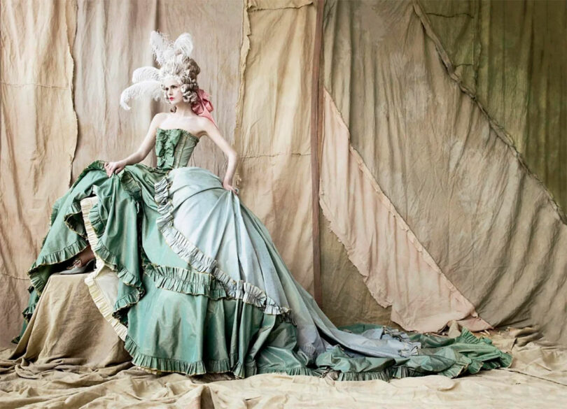 Person dressed in an elaborate green gown with a long train and feathered headdress, posed against a draped fabric background.