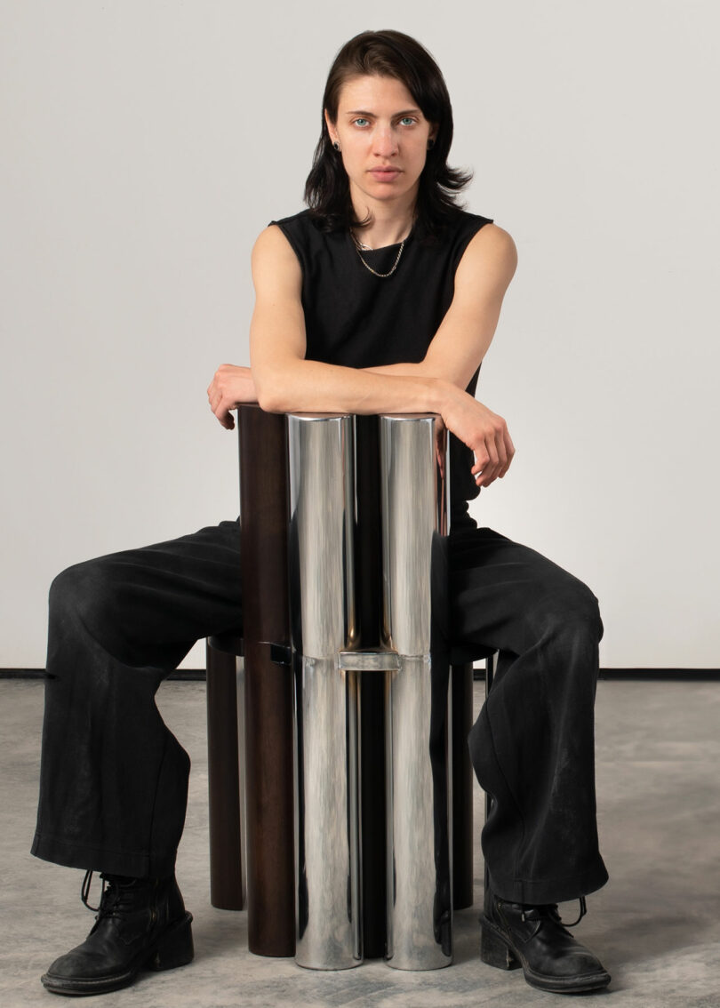 Person sitting on a cylindrical metal stool, wearing a black sleeveless top and pants, against a plain background.