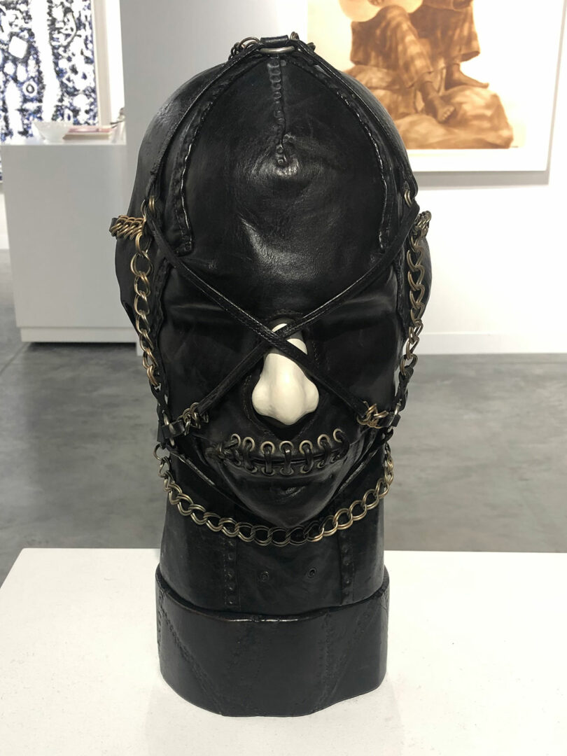 A sculpture of a black leather mask with chains and metal rings, displayed on a pedestal in a gallery setting.