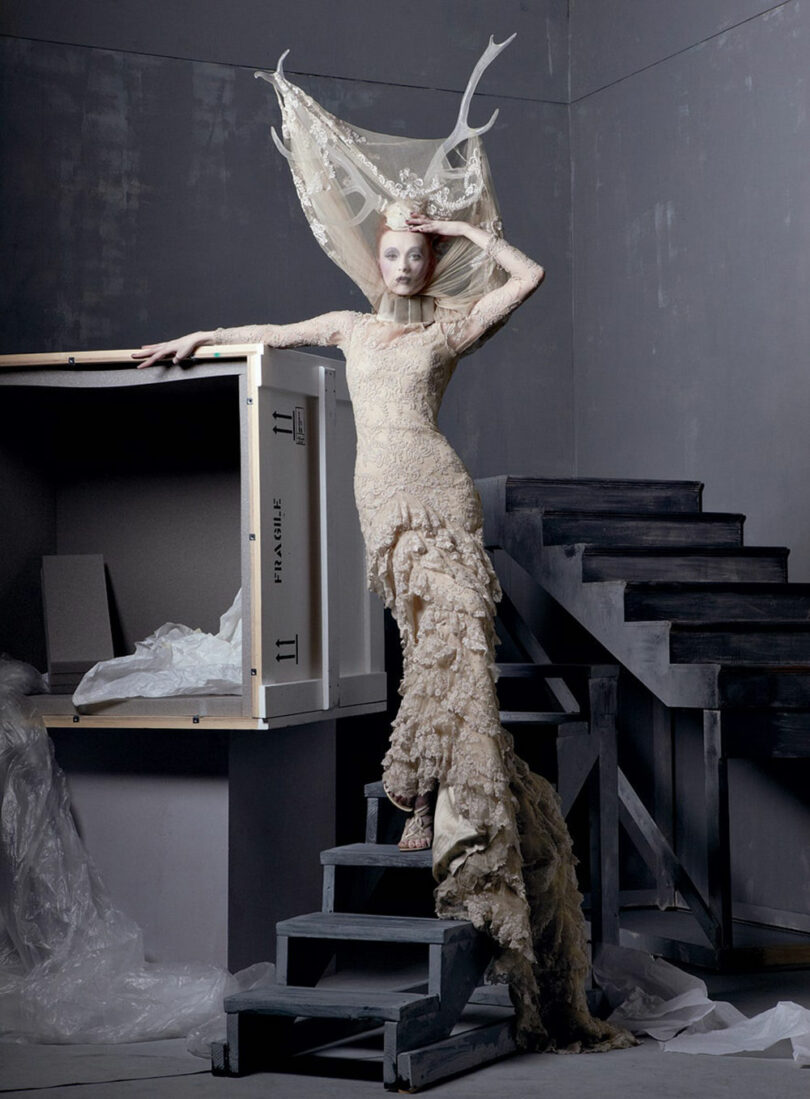 A person in an elaborate lace dress and headpiece stands on a wooden staircase next to a large package marked "FRAGILE" in a gray room.