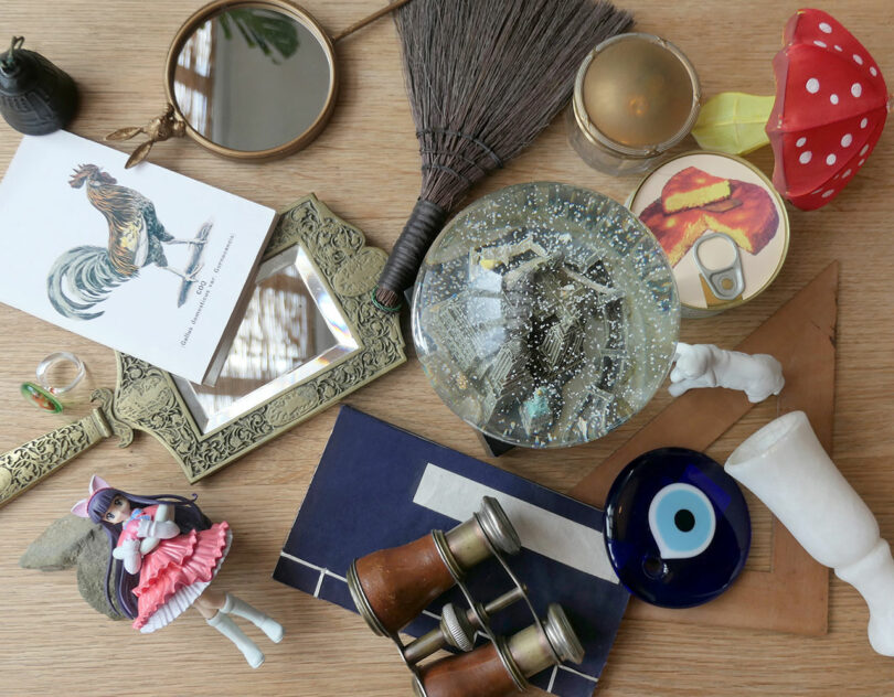 A wooden table with assorted items, including a broom, framed mirrors, binoculars, figurine, postcard, snow globe, candle, mushroom decor, evil eye, and a decorative hand.