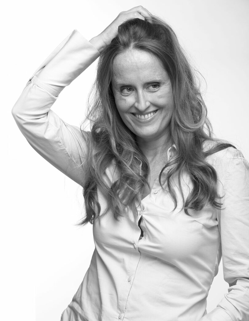 A smiling woman with long hair and a light blouse poses with one hand on her head against a plain background.