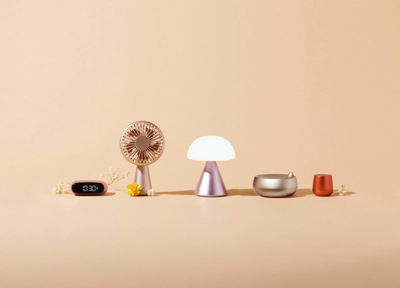 A minimalist arrangement of a digital clock, small fan, table lamp, bowl with lid, and cup against a beige background.