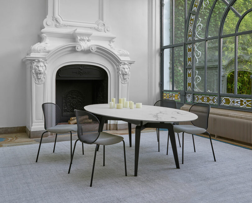 Modern dining room with an oval table, four chairs, and candles. Large window with decorative metalwork and a white ornate fireplace. Light gray carpet on the floor.