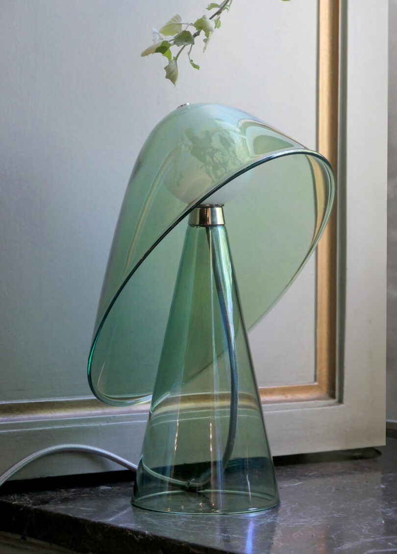 A modern glass lamp with a cone-shaped base and a dome-shaped shade is placed on a dark surface near a window. A small plant is visible in the background.