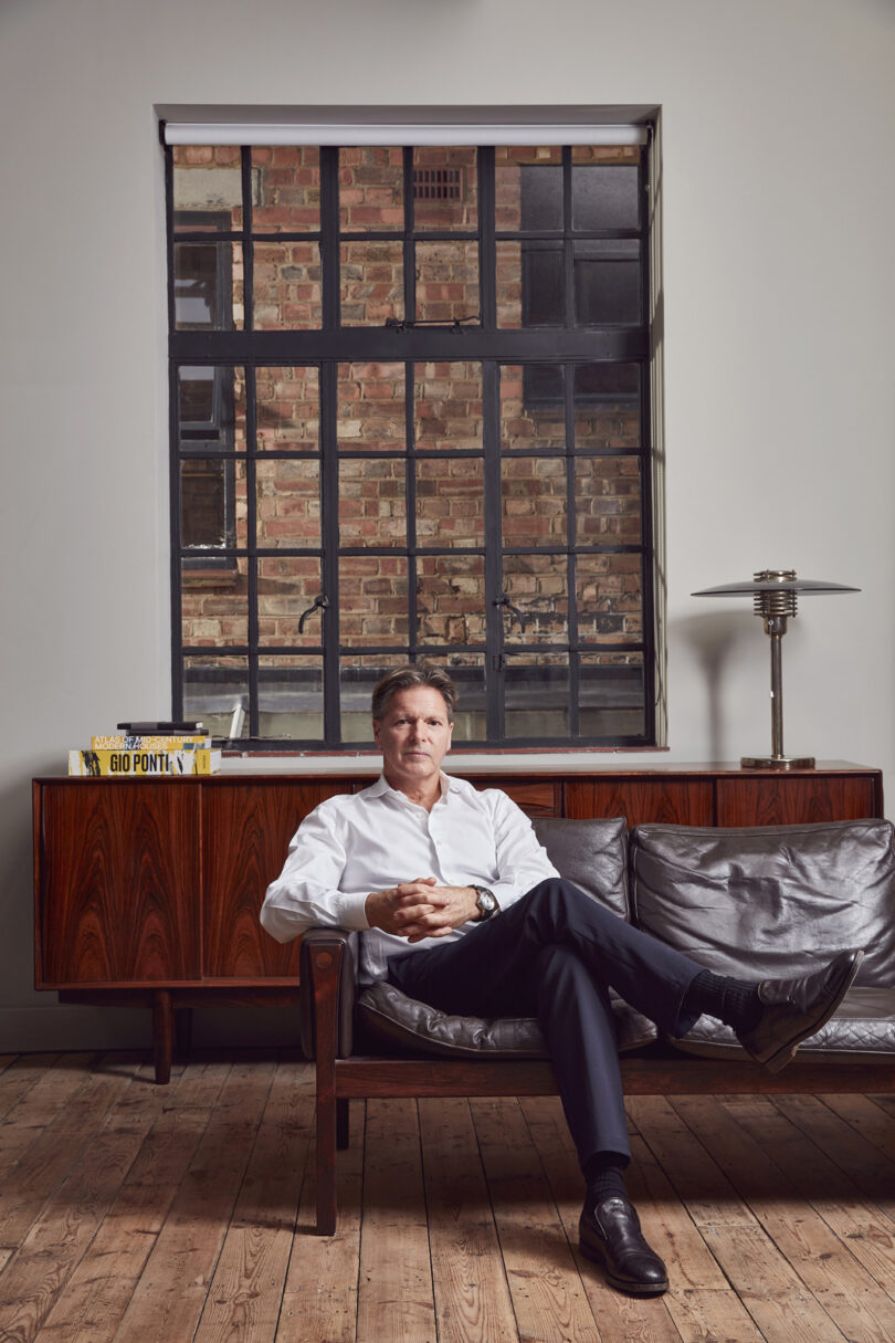 Richard Parr sits on a dark leather couch, his white shirt contrasting with the rustic charm of a large window offering a brick wall view. The wooden floors and sideboard, adorned with books and a lamp, complete the scene's cozy ambiance.