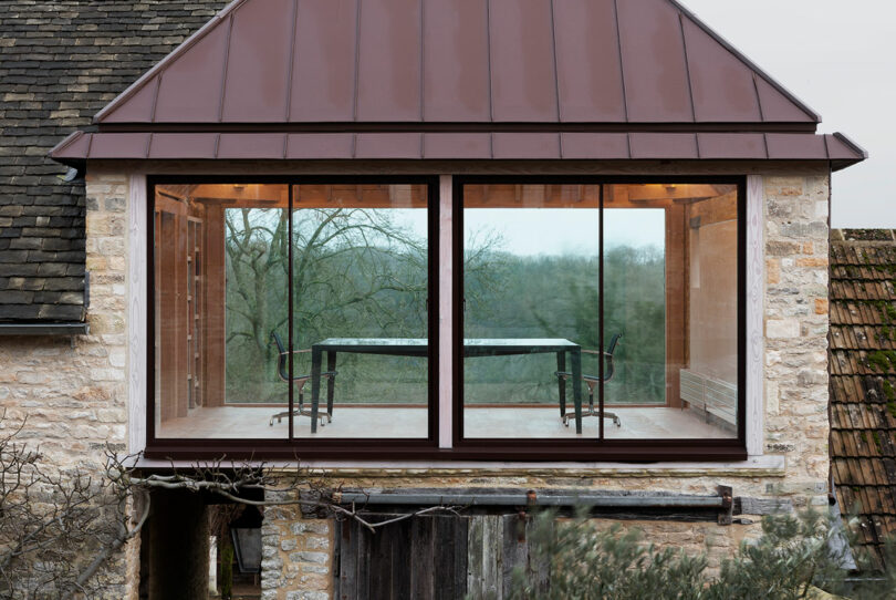 A rustic building with a modern Richard Parr-designed glass-walled room extending over the roof, featuring a table and chairs inside, overlooking a wooded landscape.