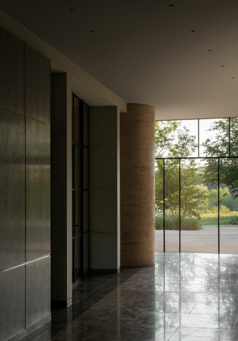 A modern interior designed by Richard Parr features large windows that welcome natural light, highlighting stone columns and dark wood paneling. Outside, the landscape is adorned with lush green trees under a clear sky.