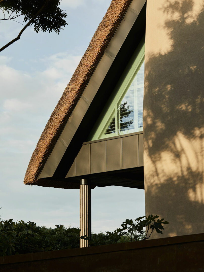 A modern building by Richard Parr, featuring a thatched roof, triangular windows, and textured walls, all partially shaded by trees.