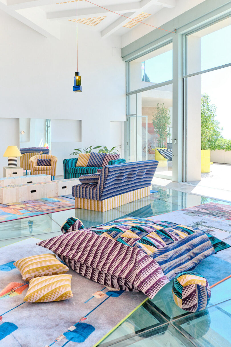 Colorful, modern living room with patterned sofas and cushions on a glass floor. Large windows and bright natural light enhance the vibrant decor