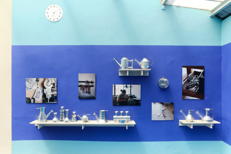 Gallery wall with blue background, displaying metal pitchers on shelves and photos. A clock is mounted above