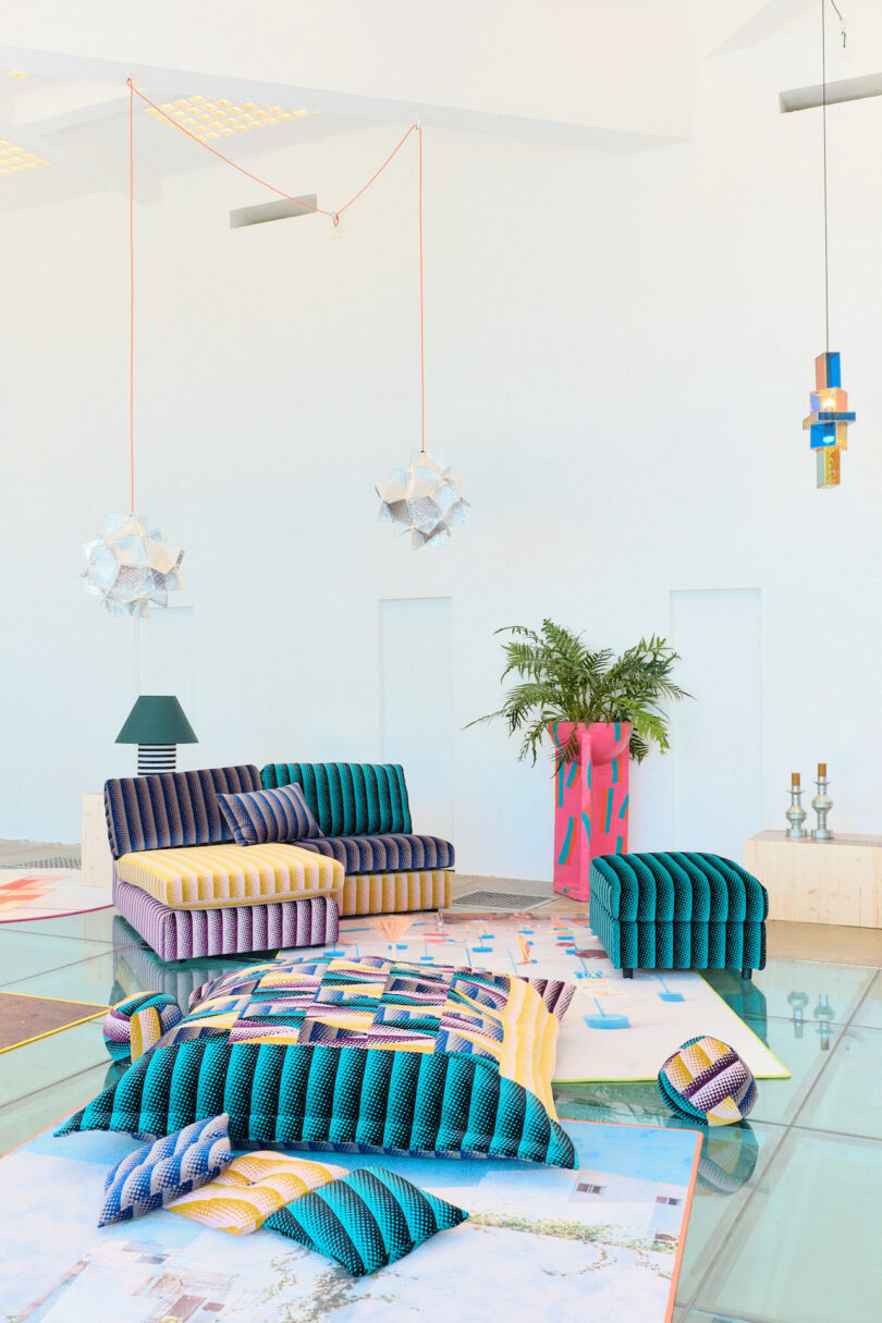 A modern living room with colorful, geometric-patterned furniture, hanging pendant lights, and a tall plant in a vibrant pot. The floor is glass, and décor is eclectic