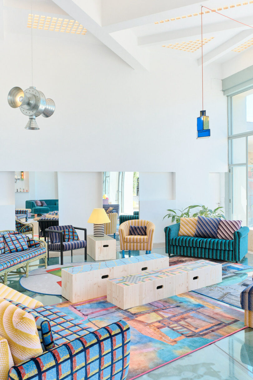 Modern living room with vibrant, patterned sofas and chairs, wooden coffee tables, colorful rugs, large windows, and unique pendant lights