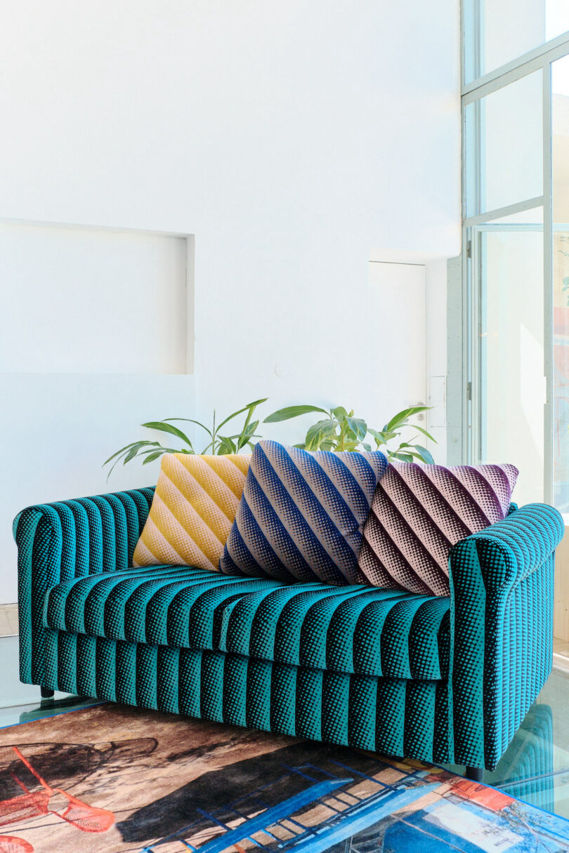 A teal upholstered sofa with three striped cushions in yellow, blue, and purple. A plant is visible behind the sofa, set against a bright white wall and large window